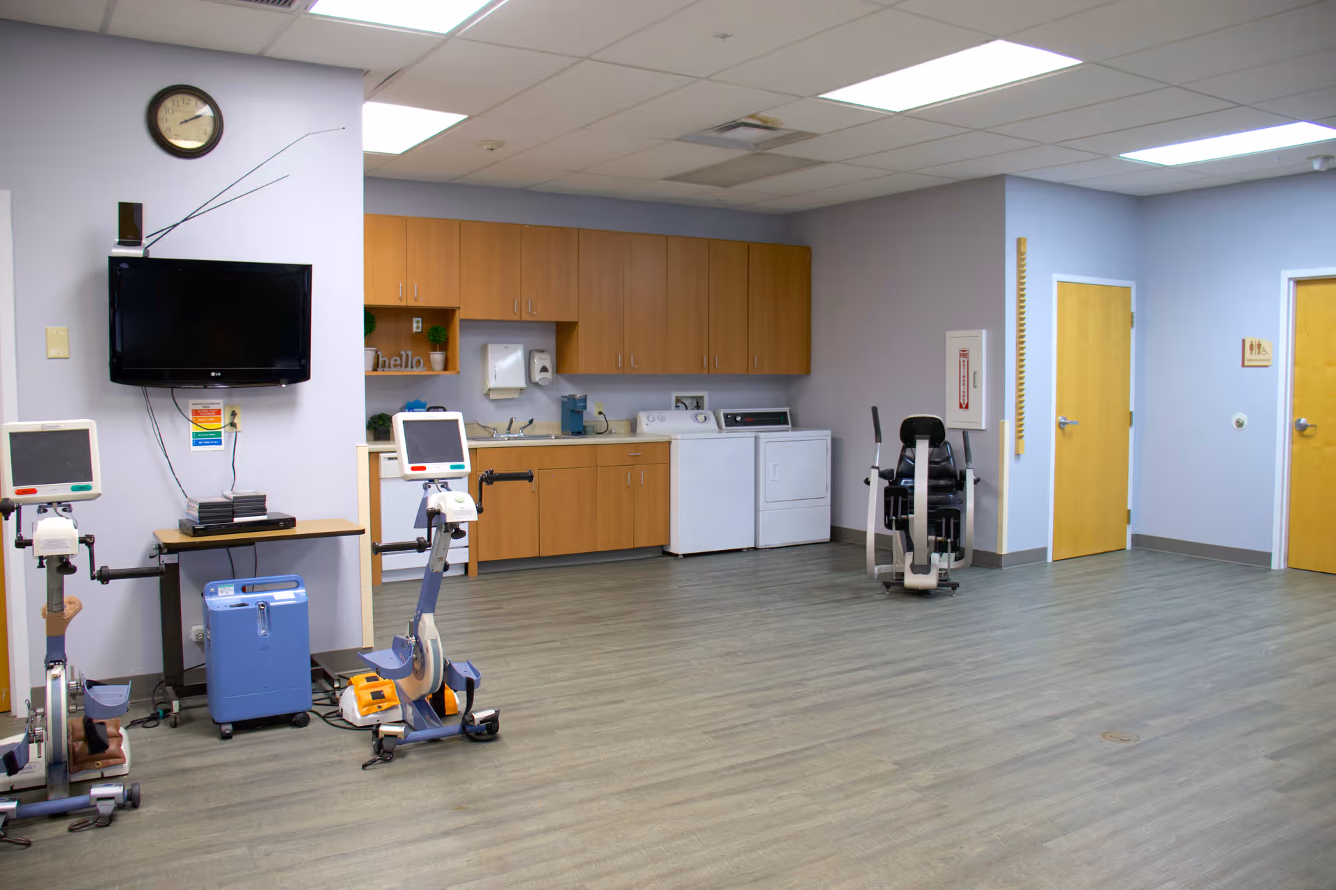 A spacious room with exercise equipment including two stationary bikes with monitors and a seated exercise machine. The room has light purple walls, wood laminate flooring, and a ceiling with fluorescent lights. There is a wall-mounted TV, a clock, wooden cabinets, a sink, a washing machine, and a dryer along one wall. Two closed wooden doors are visible on the right side of the room.