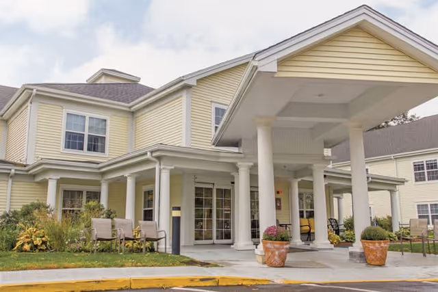 Exterior view of a senior living facility building with yellow siding, white columns, and a covered entrance. There are chairs and plants near the entrance, and the sky is partly cloudy.