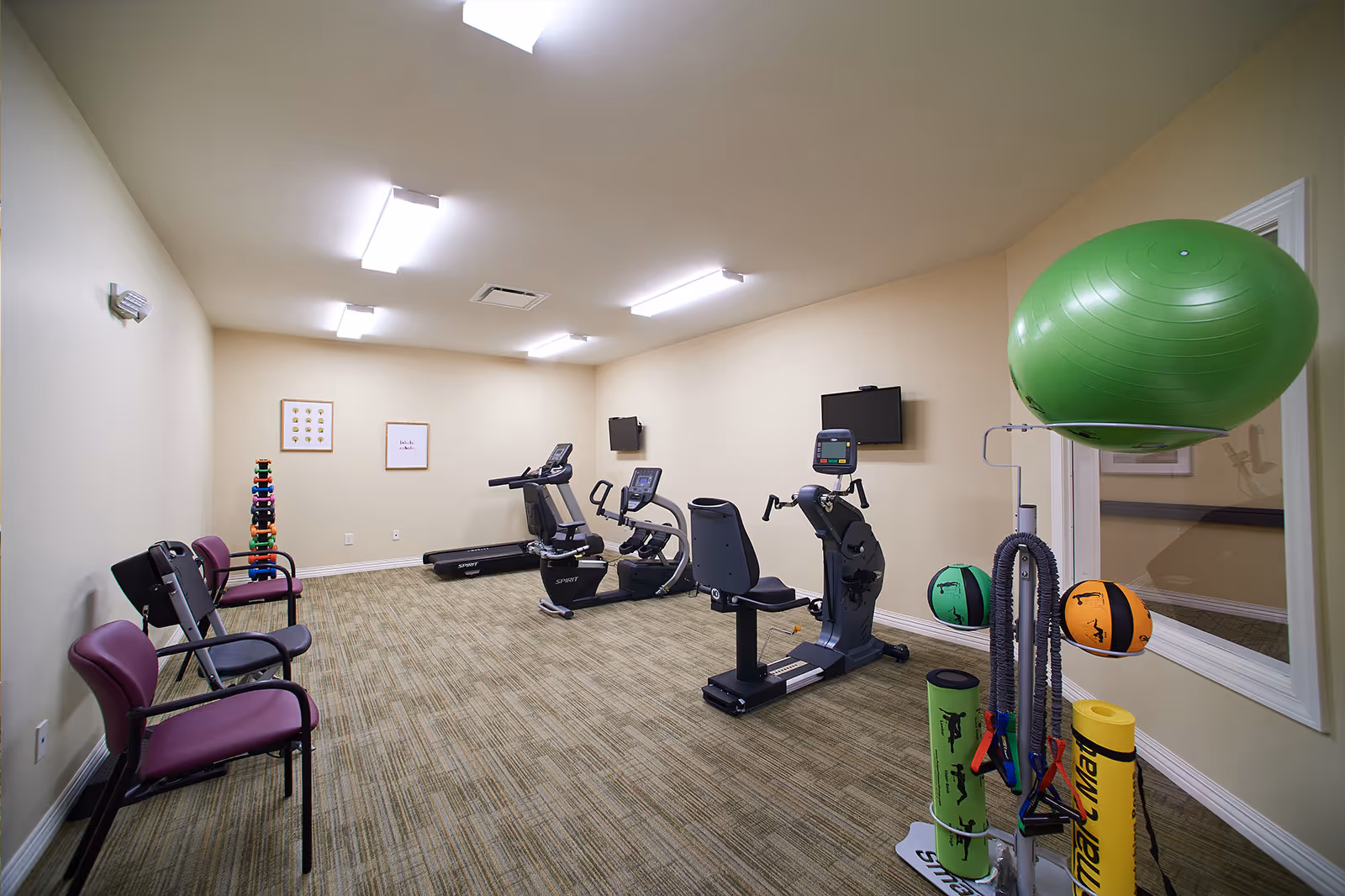A small fitness room with exercise equipment including two recumbent bikes, a treadmill, and various exercise accessories such as a large green exercise ball, resistance bands, yoga mats, and medicine balls. There are three chairs along one wall and two framed pictures hanging on the far wall. The room has beige walls, carpeted flooring, and bright overhead lighting.