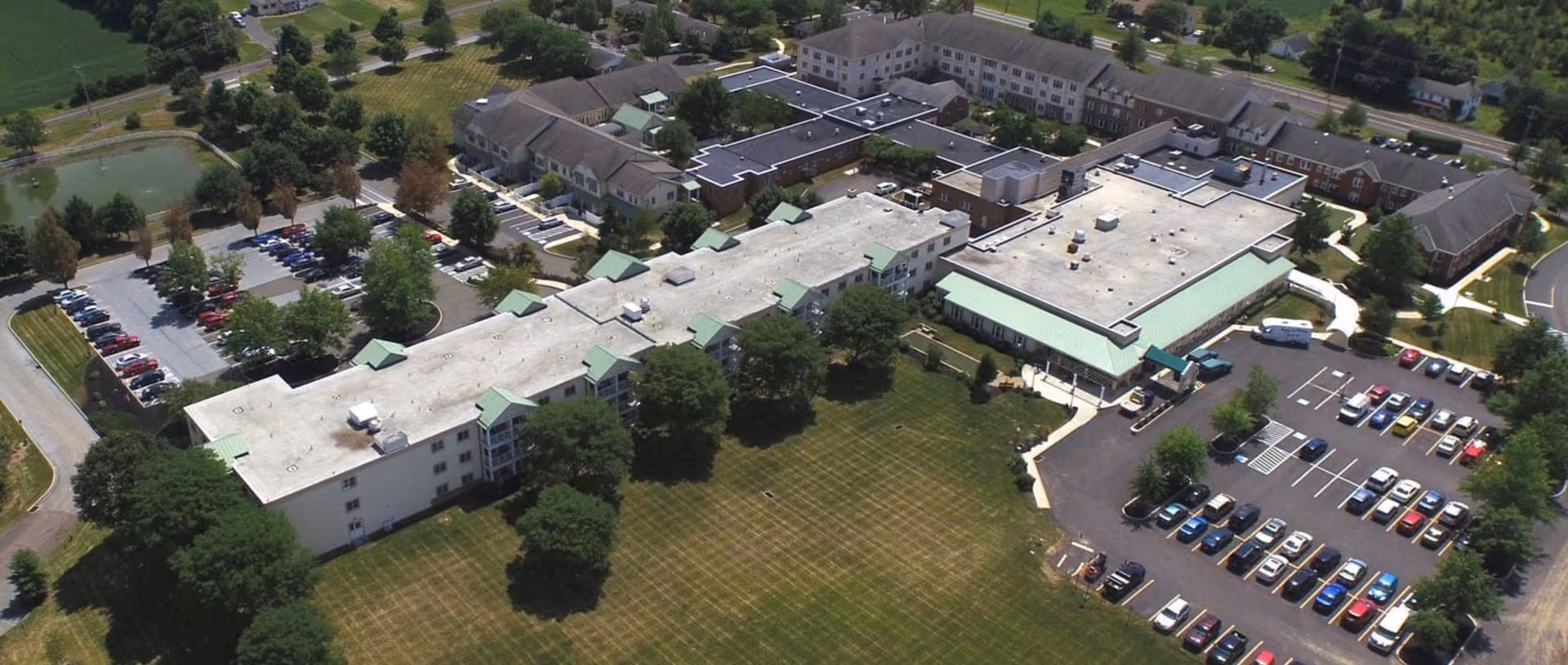 Aerial view of Frederick Living facility showing multiple connected buildings with flat roofs, surrounded by green lawns, trees, parking lots filled with cars, and nearby roads.