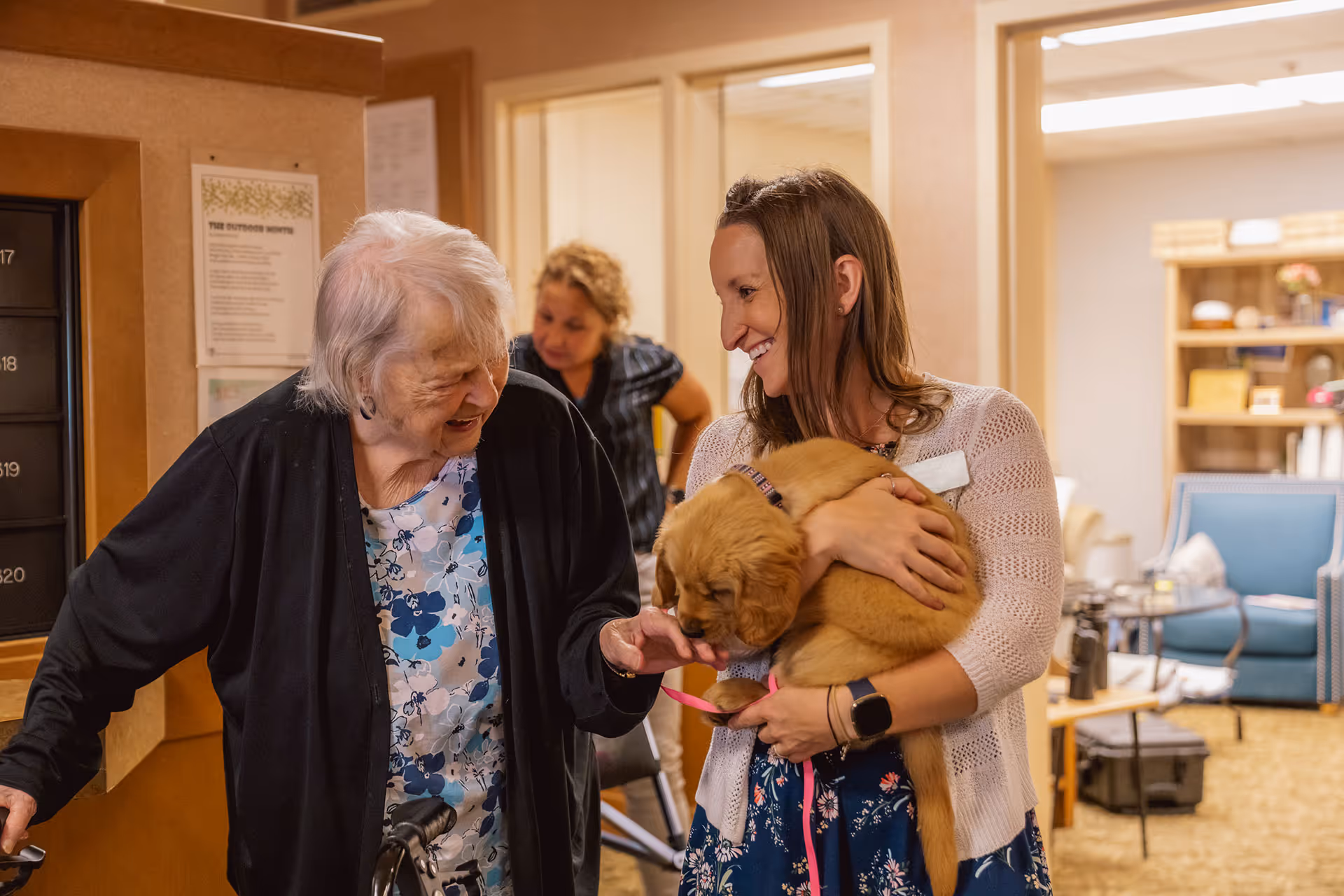 An elderly woman with a walker smiles and interacts with a younger woman holding a golden retriever puppy inside a senior living facility. Another woman is seen in the background. The setting appears warm and welcoming with comfortable seating and soft lighting.