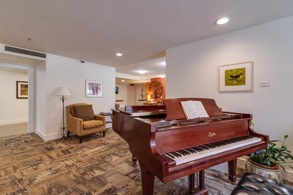 Interior view of a senior living facility common area featuring a polished wooden grand piano with sheet music on the stand, a cushioned armchair next to a floor lamp, framed artwork on the walls, and patterned carpet flooring. The space is well-lit with ceiling lights and has a cozy, welcoming atmosphere.
