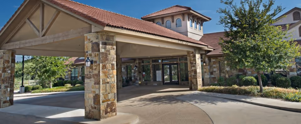 Exterior view of a senior living facility entrance with a covered driveway supported by stone pillars, a tiled roof, and surrounding landscaping including trees and shrubs under a clear blue sky.