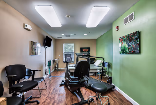 Small fitness/therapy room with a recumbent bike, treadmill, parallel bars, chairs, and wall art.