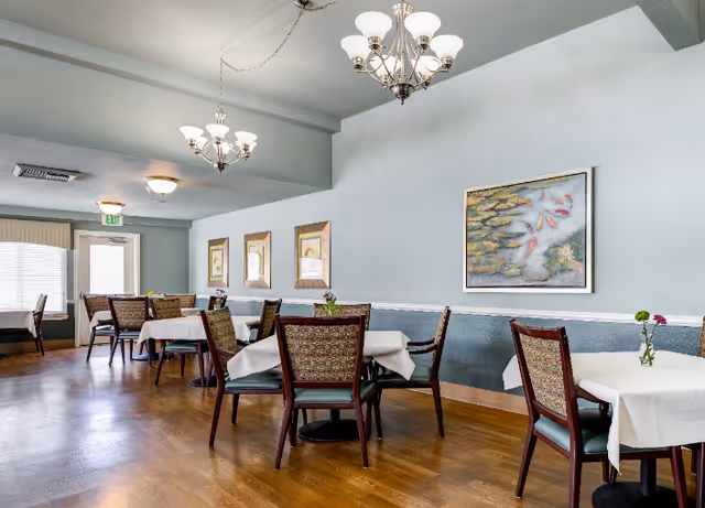 Bright dining room with multiple small tables draped in white cloths, upholstered chairs, chandeliers, framed artwork, and hardwood floors.