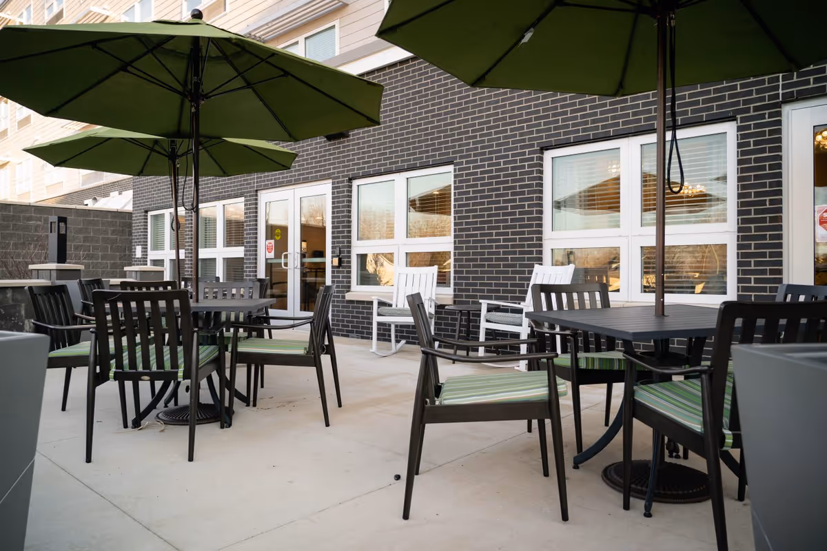 Outdoor patio with tables, green umbrellas, and chairs outside a brick-faced senior living building.