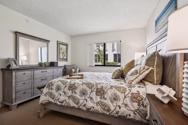 A well-lit bedroom with a large bed featuring a patterned bedspread and multiple pillows. There is a wooden dresser with a large mirror on the left side, a framed picture on the wall, and a window with a view of greenery outside. A bedside table with a lamp and decorative item is on the right side of the bed.
