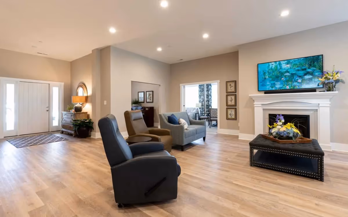 Bright communal living room with recliner chairs, a sofa, a fireplace topped by a wall-mounted TV, and a coffee table on wood flooring.