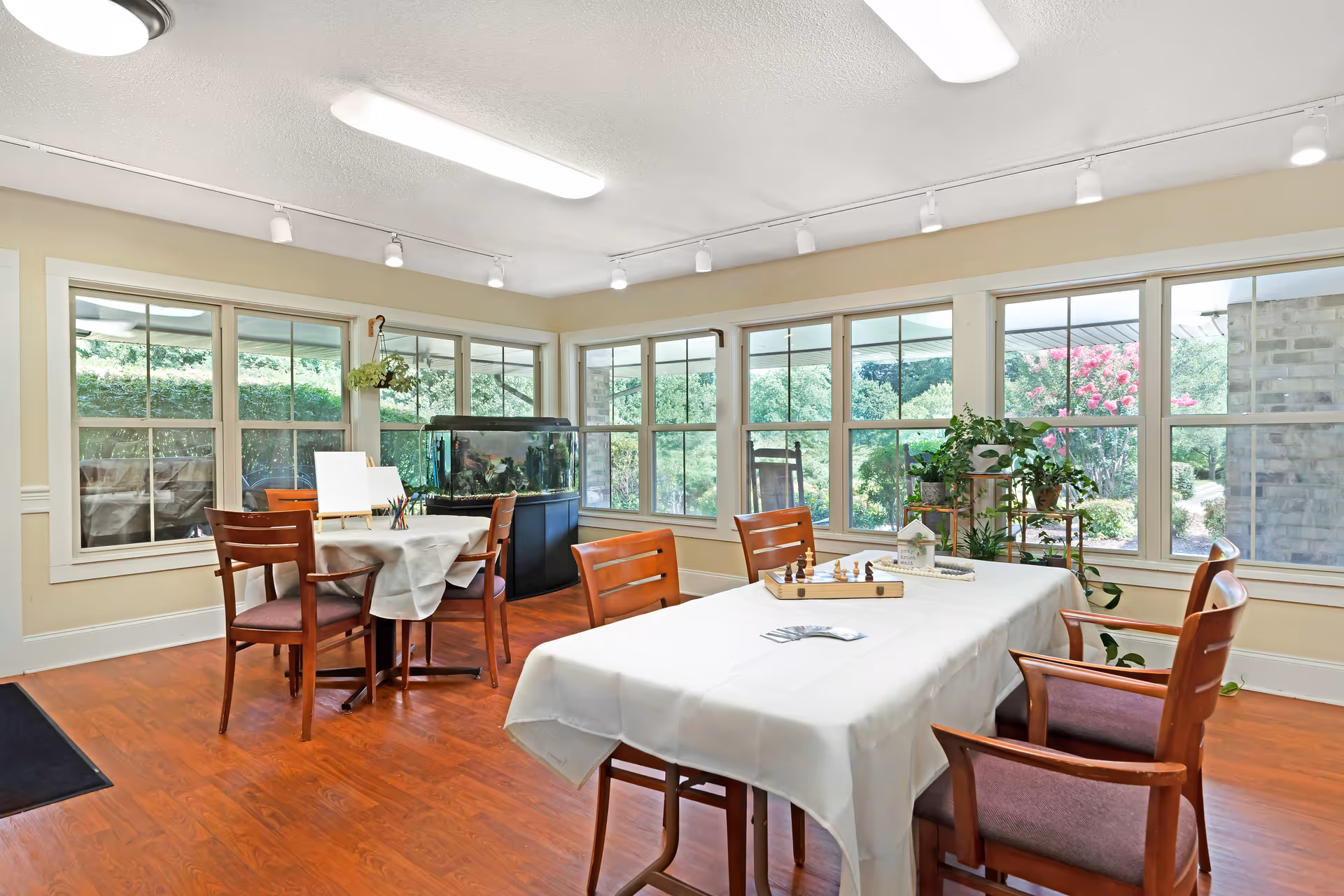 A bright activity room with large windows showing greenery outside. The room has wooden floors and several wooden chairs around tables covered with white tablecloths. One table has a chessboard and cards, while another has art supplies and small canvases. There is a large fish tank against the wall and several potted plants near the windows.