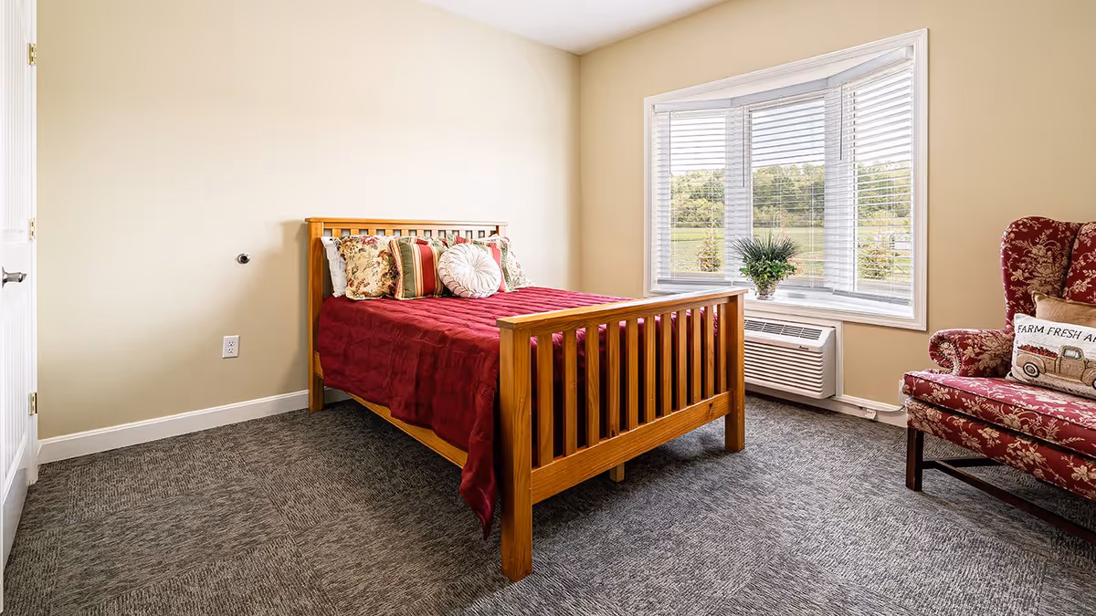 Bright bedroom with a wooden bed dressed in a red coverlet, a patterned armchair, and a bay window overlooking a lawn.