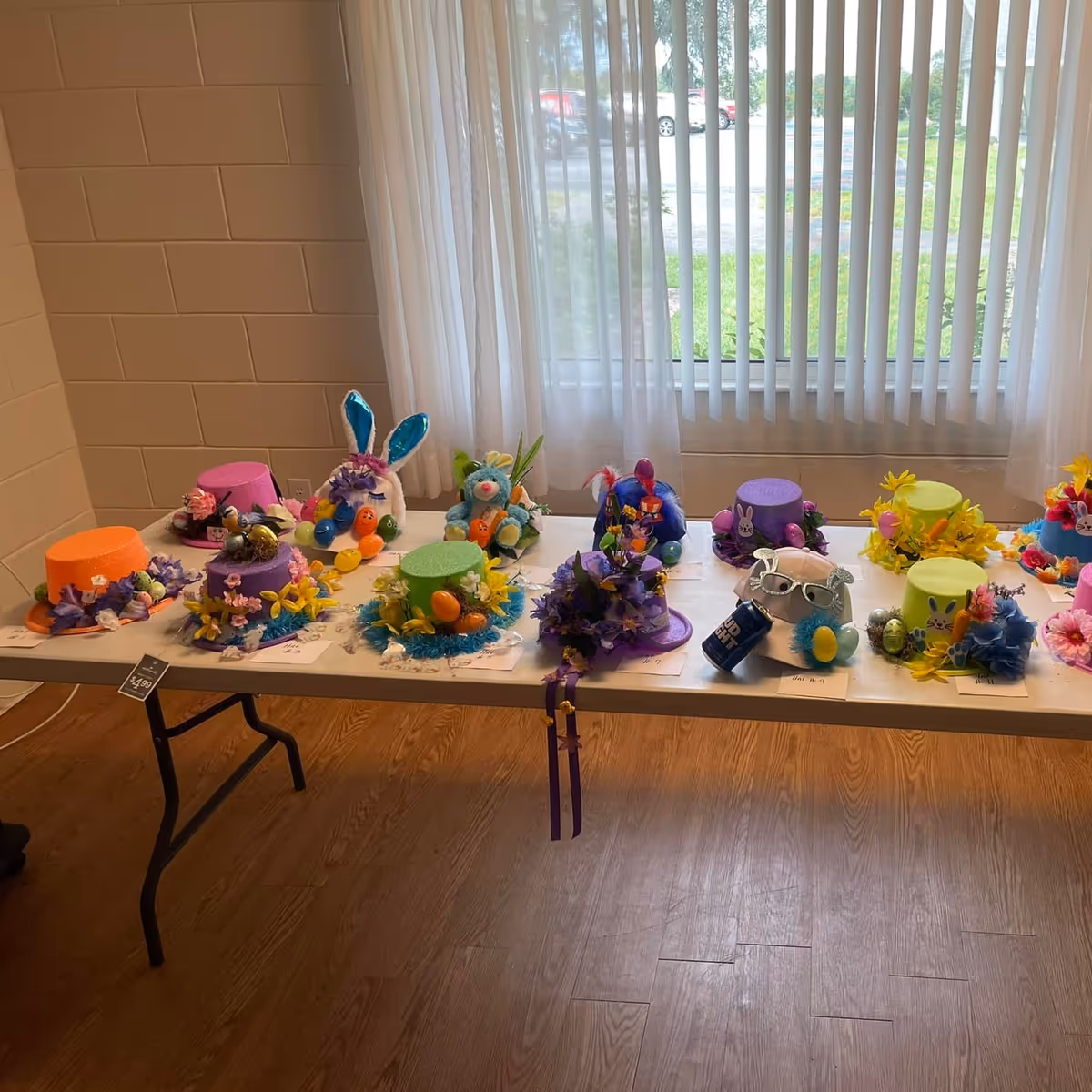 A table inside a room displaying a variety of colorful decorated hats, some with Easter-themed decorations like bunny ears, eggs, and flowers. The table is positioned in front of a window with vertical blinds, and the room has wooden flooring and beige brick walls.