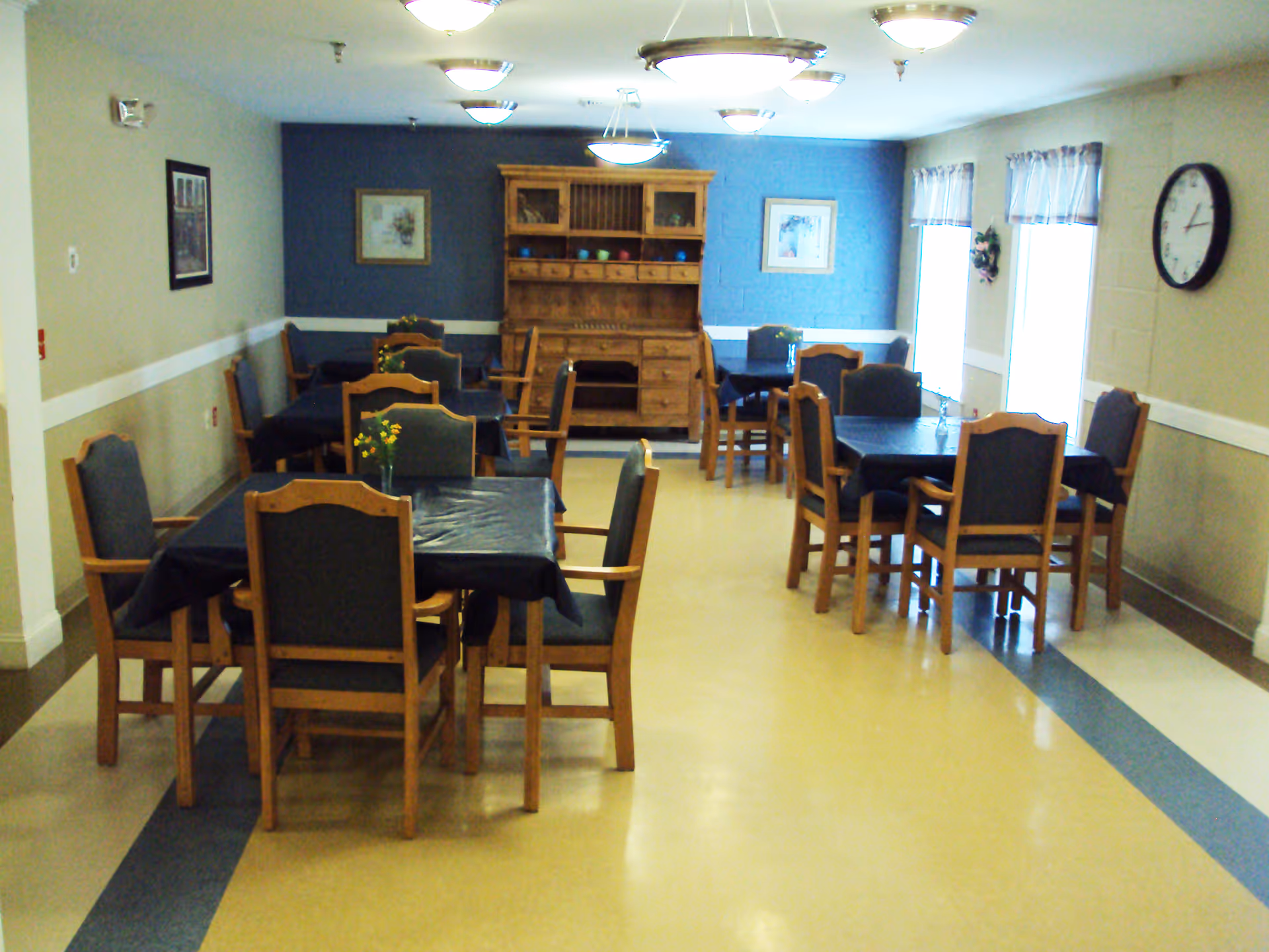 A dining room with several wooden tables and chairs arranged neatly. Each table is covered with a dark tablecloth and has a small vase with yellow flowers. The room has a light yellow floor with blue accents, beige walls with a white chair rail, and a blue accent wall at the far end. There are framed pictures on the walls, a wooden hutch with decorative items, windows with curtains on the right side, and a clock on the wall.