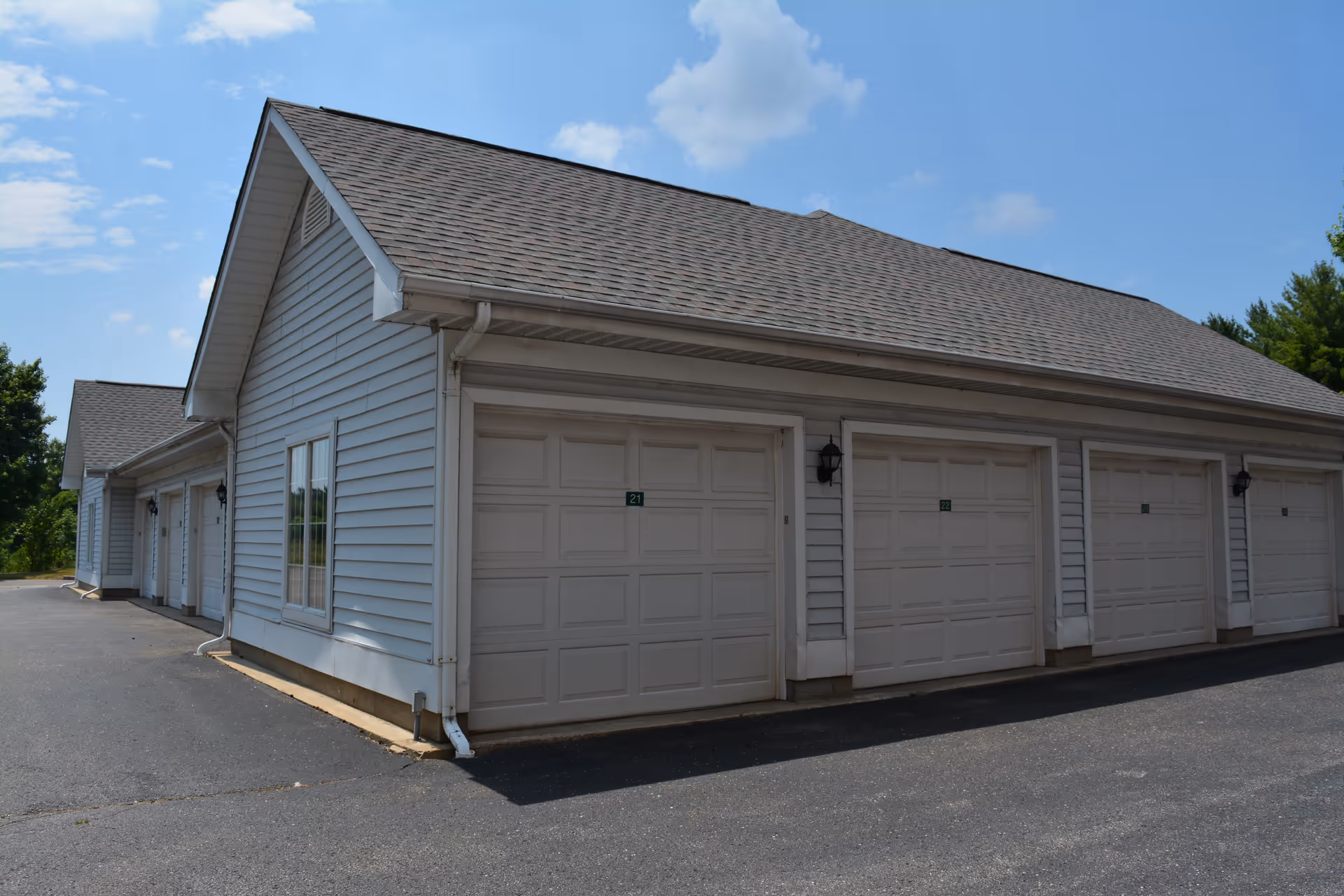 Exterior view of a row of attached garages with white doors and gray siding under a shingled roof on a sunny day with a partly cloudy sky.