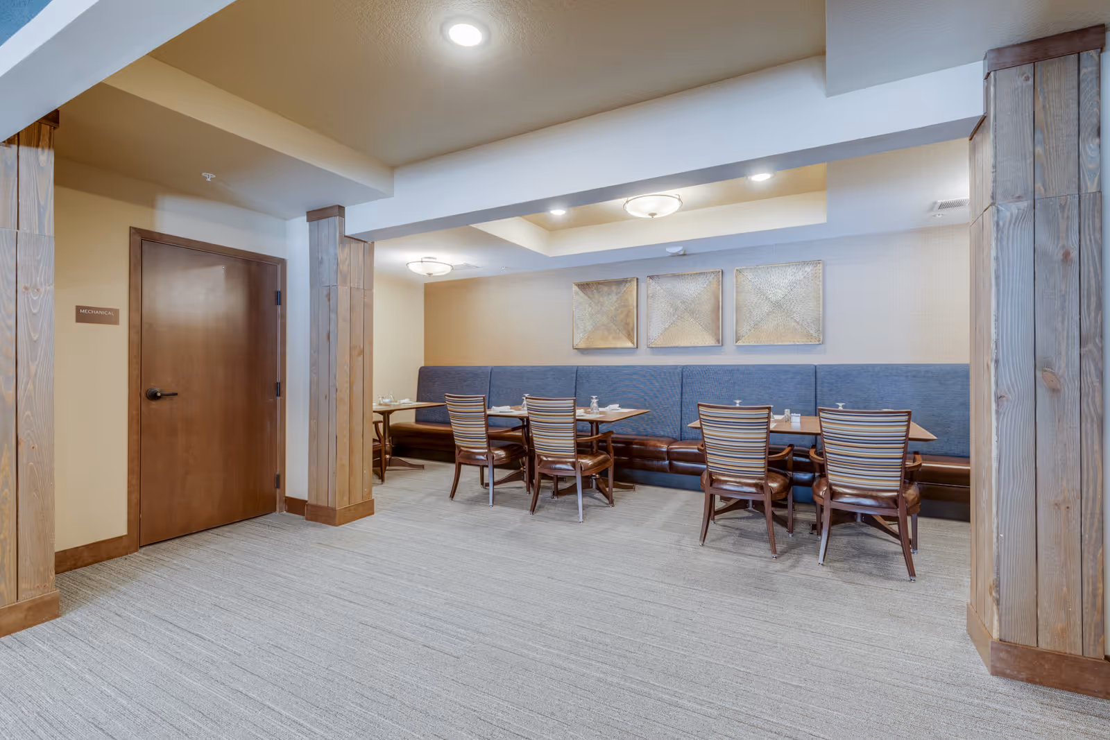 A dining area with wooden tables and chairs arranged along a blue cushioned bench against the wall. The room features beige walls, carpeted flooring, wooden pillars, and ceiling lights. There is a closed wooden door on the left side with a small sign next to it.