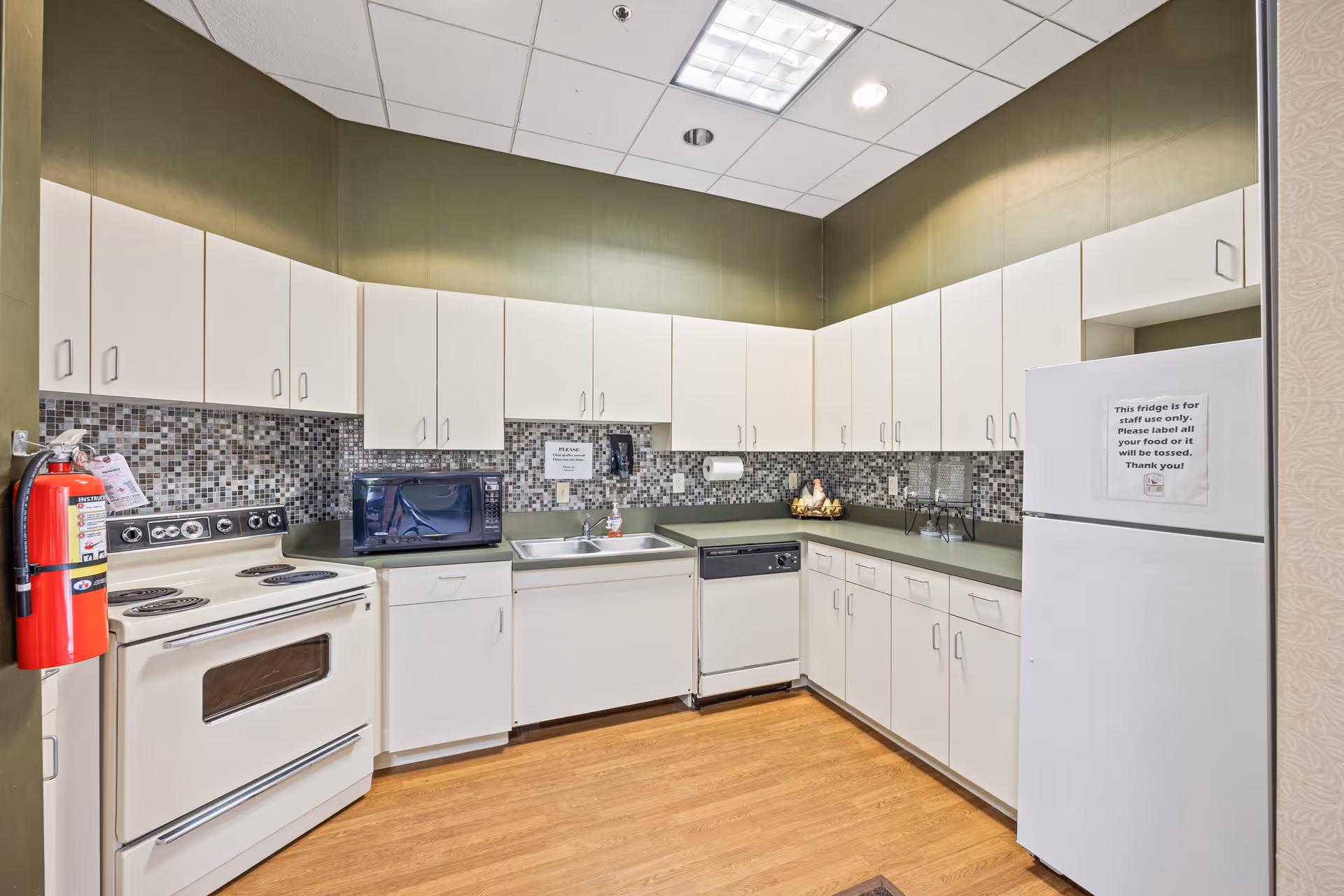 A clean and well-lit kitchen with white cabinets, a white stove, a microwave, a dishwasher, a double sink, and a white refrigerator. The backsplash features small mosaic tiles in neutral tones, and the floor is wooden. A fire extinguisher is mounted on the left wall. There are signs on the wall and refrigerator with instructions for staff use.