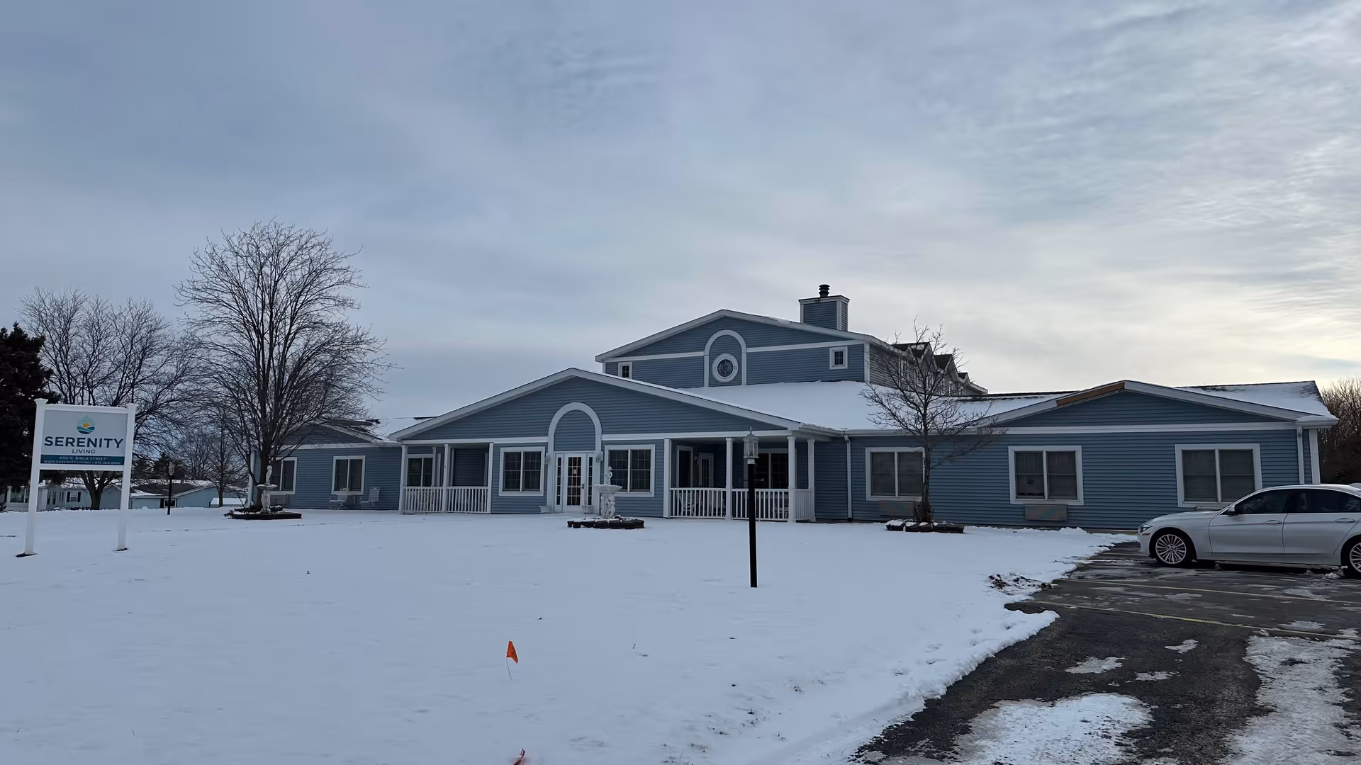 Exterior view of Serenity Living facility during winter with snow covering the ground. The building is light blue with white trim and has multiple sections with a central entrance. There are leafless trees and a parked white car on the right side. A sign with the Serenity Living logo is visible on the left side of the image.