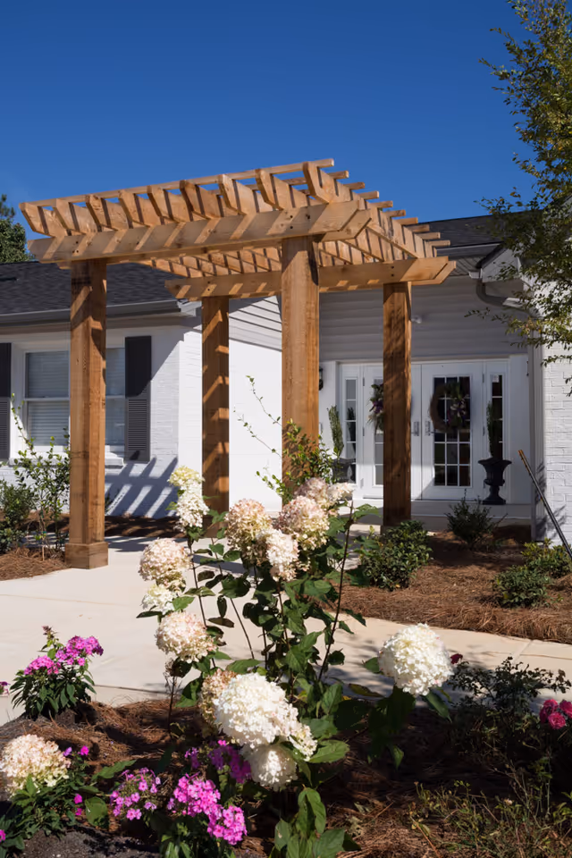 Outdoor garden area with blooming white and pink flowers in front of a wooden pergola structure. Behind the pergola is the entrance to a building with white walls, black shutters, and double glass doors decorated with wreaths. The sky is clear and blue.