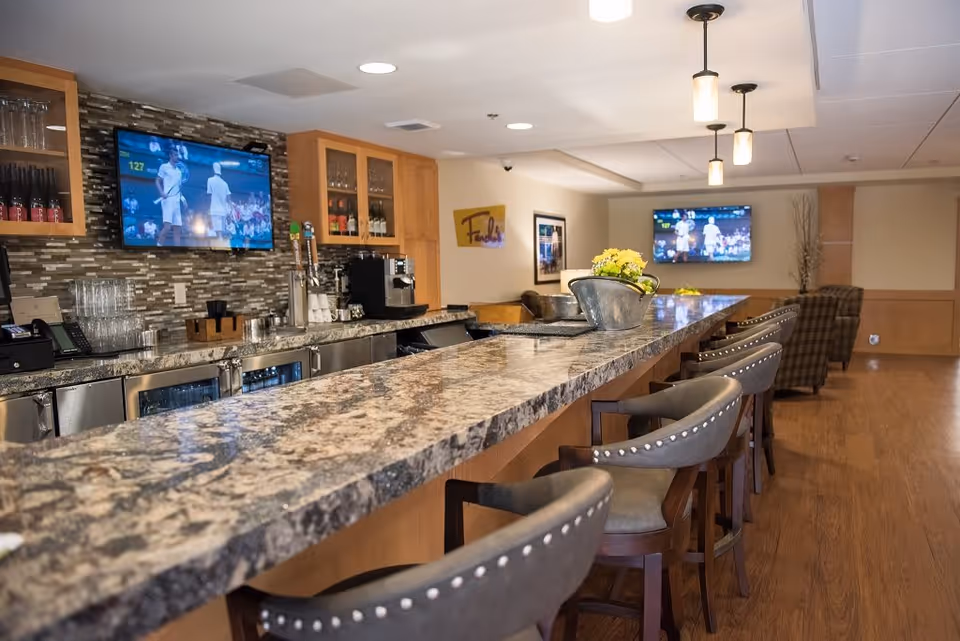Interior view of a senior living facility's bar area with a long granite countertop, several bar stools with studded backs, a tiled backsplash, and two wall-mounted televisions showing a tennis match. The space has wooden flooring, pendant lighting, and a seating area with armchairs in the background.