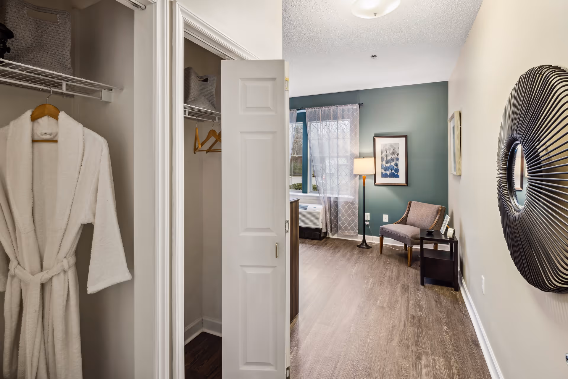 View of a cozy interior room with a partially open closet door showing a white bathrobe hanging inside. The room features wooden flooring, a green accent wall with a framed picture, a window with sheer curtains, a floor lamp, a cushioned chair, a small side table, and a decorative round mirror on the opposite wall.