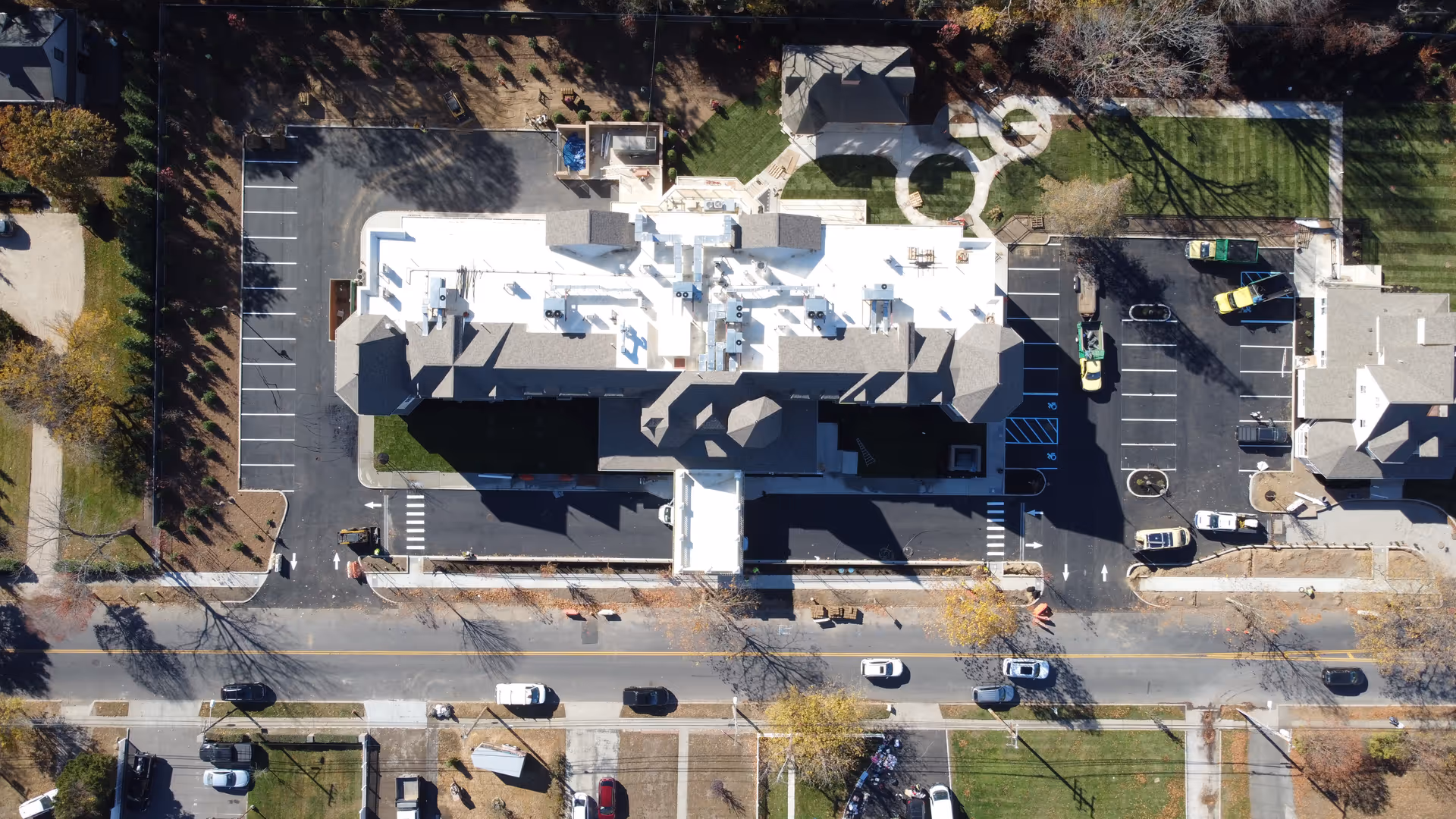 Aerial view of a senior living facility named Netherbay at Bay Shore, showing the building with a white roof, surrounding parking lots with several parked vehicles, landscaped green areas, and a road with cars in front of the facility.