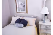 A neatly made bed with white linens, two dark blue pillows, and a rolled gray towel placed on top. The bed has a white headboard and is positioned against a light-colored wall with a framed picture hanging above it. To the right of the bed is a white nightstand with drawers, a white table lamp, and some small items on top.