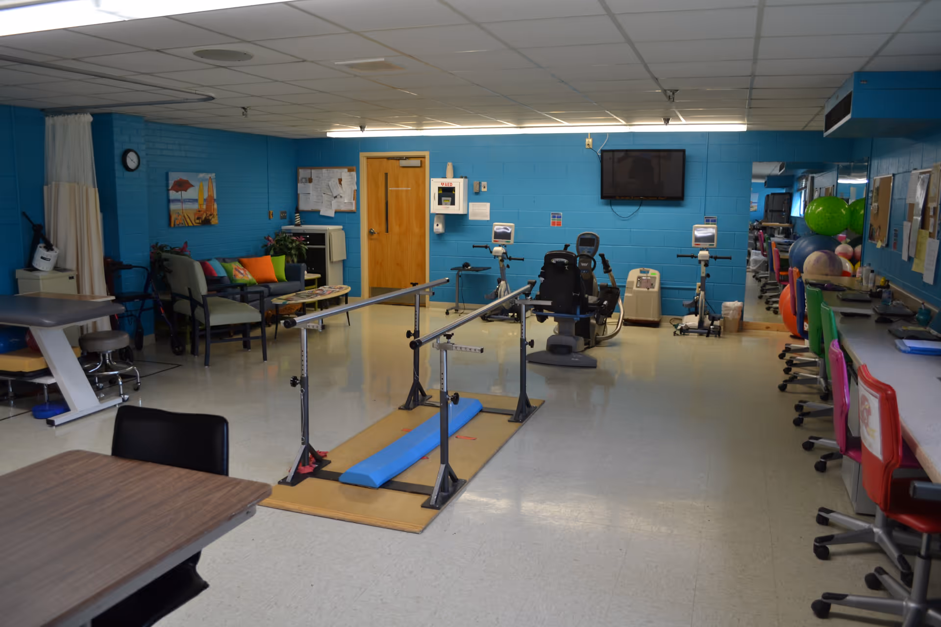 Interior view of a therapy room with blue walls, featuring parallel bars for walking exercises in the center, exercise bikes and equipment against the back wall, a seating area with colorful pillows on the left, and a row of chairs along a counter on the right. A wall-mounted TV is visible above the exercise equipment.