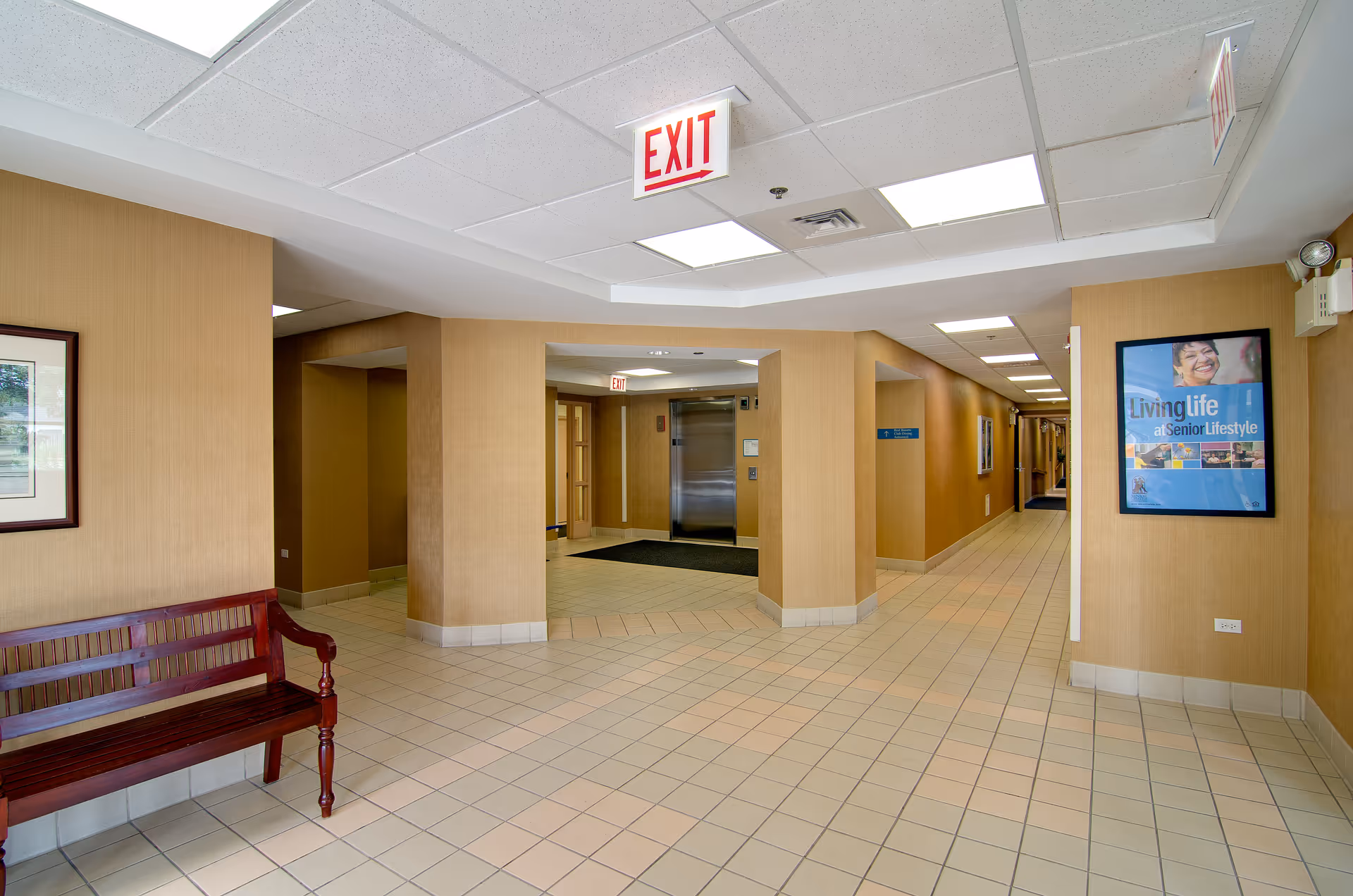 Interior view of a senior living facility hallway with beige walls and tiled floor. There is a wooden bench on the left side and an elevator straight ahead. A framed poster on the right wall reads 'Living Life at Senior Lifestyle.' An exit sign hangs from the ceiling.