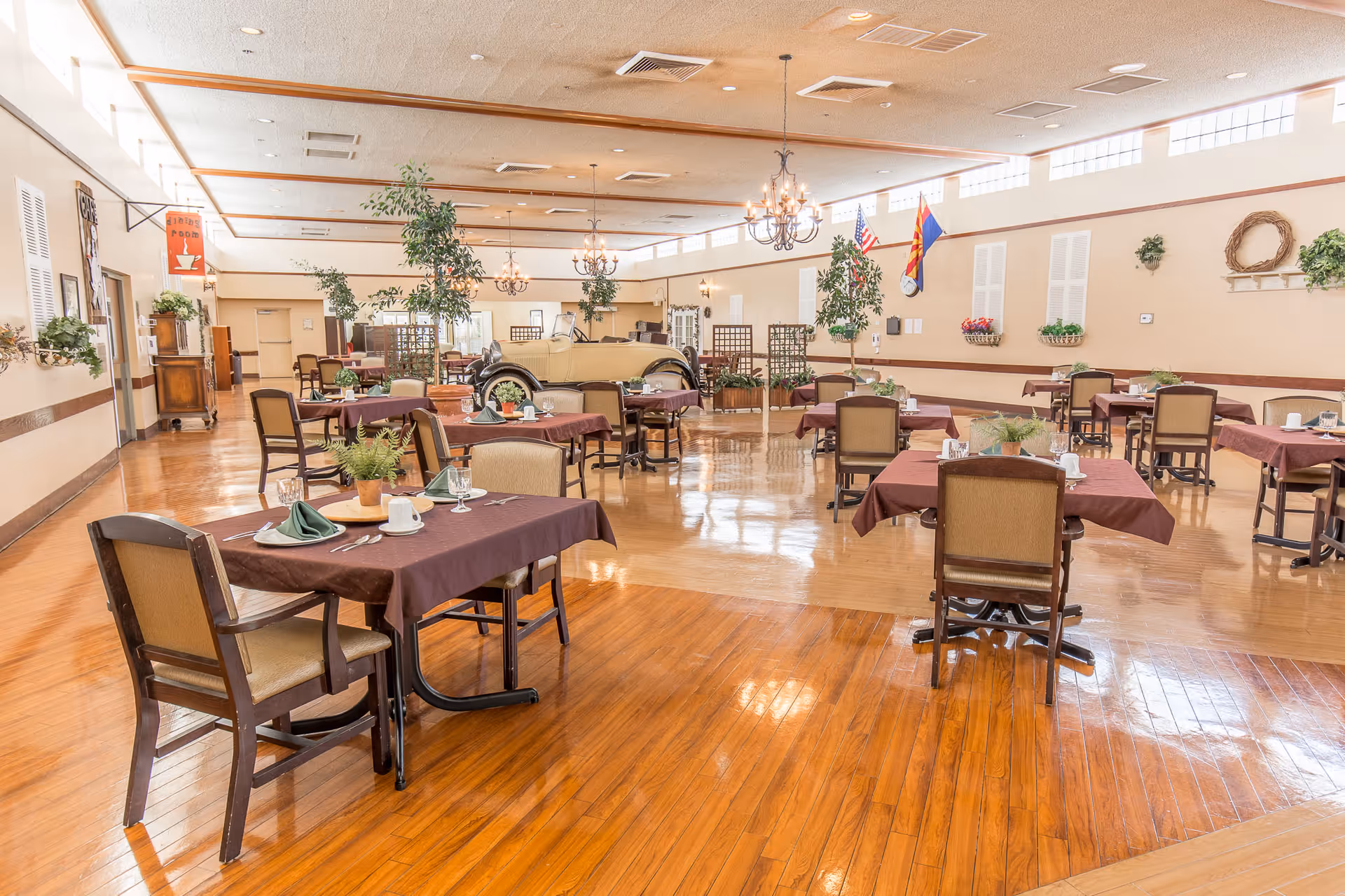 A spacious dining room with multiple tables covered in brown tablecloths, each set with plates, cups, and green napkins. The room features wooden floors, chandeliers hanging from the ceiling, several potted plants, and flags on the far wall. Large windows near the ceiling allow natural light to fill the space.