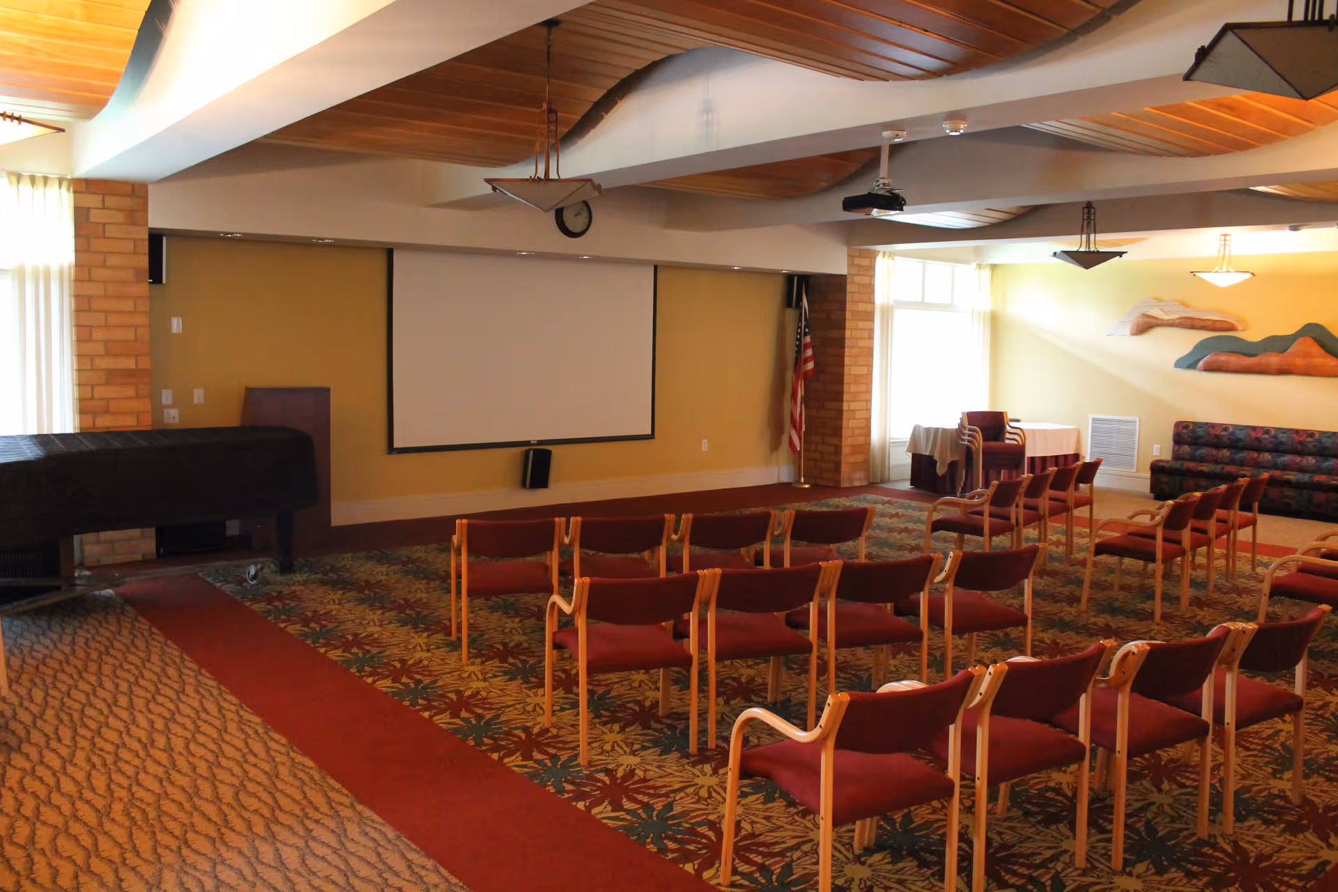 A meeting or presentation room with rows of red cushioned chairs facing a large white projection screen mounted on a beige wall. The room has a patterned carpet with floral designs, a covered piano on the left side, an American flag in the corner, and a colorful abstract wall decoration on the right. There are windows letting in natural light and ceiling lights providing additional illumination.