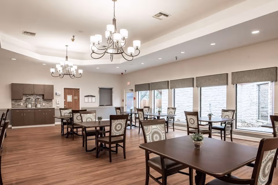 A bright dining room with several wooden tables and chairs arranged neatly. Each table has a small potted plant as a centerpiece. The room features large windows with gray valances, allowing natural light to enter. The floor is wooden, and the ceiling has two elegant chandeliers with multiple lights. In the background, there is a counter area with cabinets and two beverage dispensers.