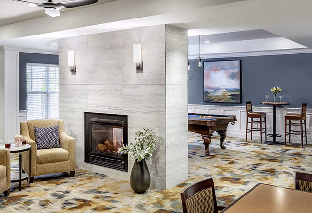 A cozy senior living common area featuring a modern fireplace set in a light stone wall with two wall sconces above it. To the left, there is a comfortable armchair with a patterned pillow and a small side table. In the background, a pool table is visible along with a high table and two chairs near a blue-gray wall adorned with a large abstract painting. The room has a patterned carpet and a ceiling fan above.