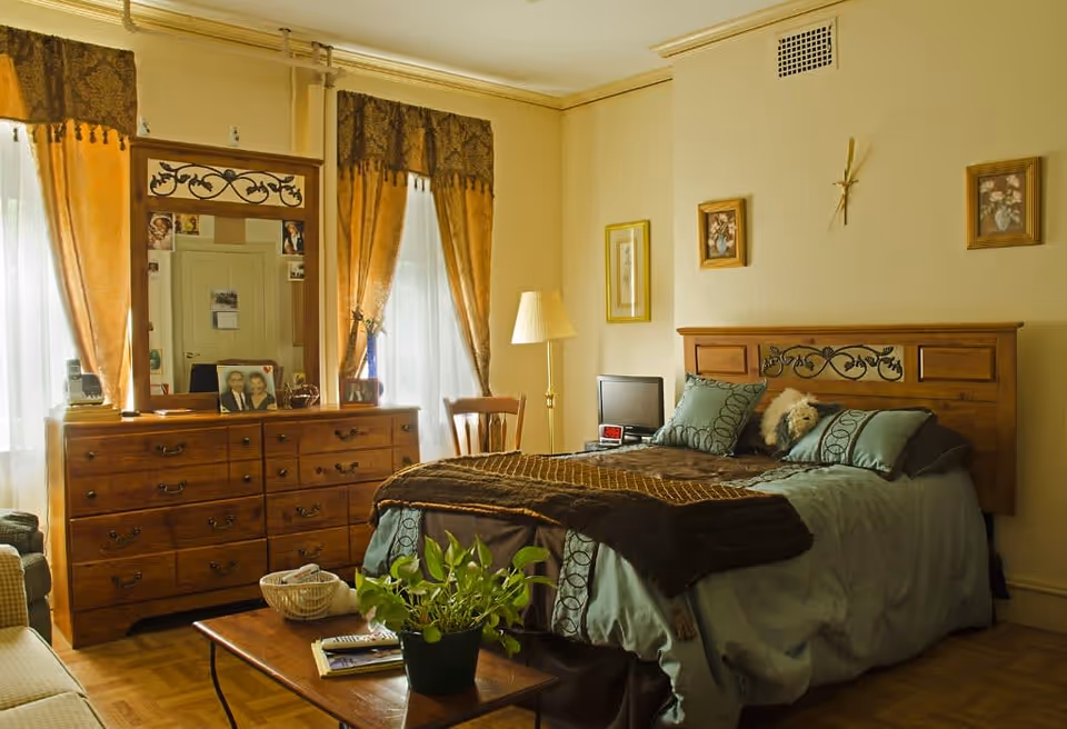 A cozy bedroom with a wooden bed featuring decorative carvings, blue and brown bedding, and a stuffed animal on the pillows. There is a wooden dresser with a large mirror, family photos, and various items on top. The room has two windows with sheer white curtains and brown drapes, a wooden chair, a floor lamp, framed artwork on the walls, and a coffee table with a green potted plant and reading materials.