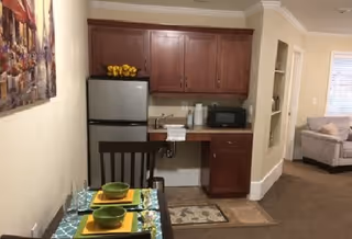 A small kitchenette area with a refrigerator, microwave, sink, and wooden cabinets. In front of the kitchenette is a dining table set for two with green bowls, yellow placemats, and glasses. To the right, there is a partial view of a living room with a beige sofa and a window with blinds.