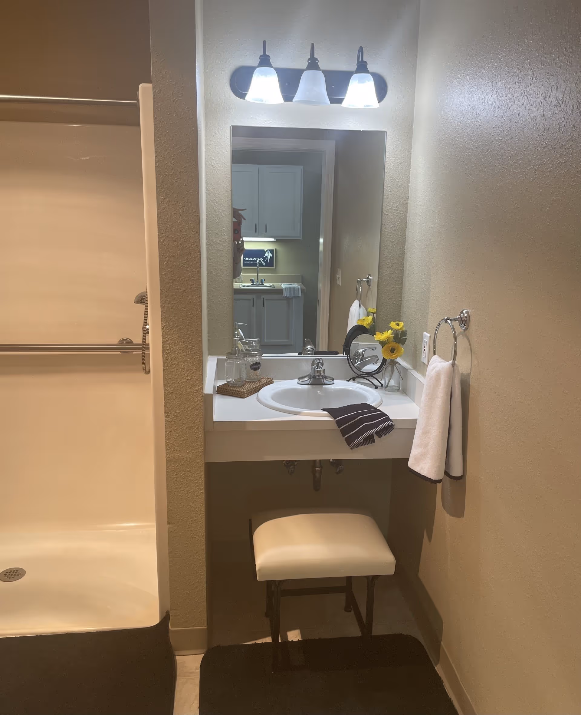 A bathroom vanity area with a white sink, a mirror above it, and three light fixtures on top. A small cushioned stool is placed under the sink. To the left, there is a shower stall with a grab bar and a handheld showerhead. A towel ring with a white towel is mounted on the wall to the right of the sink. On the vanity countertop, there are decorative items including a small vase with yellow flowers, a glass jar, and a folded dark towel.