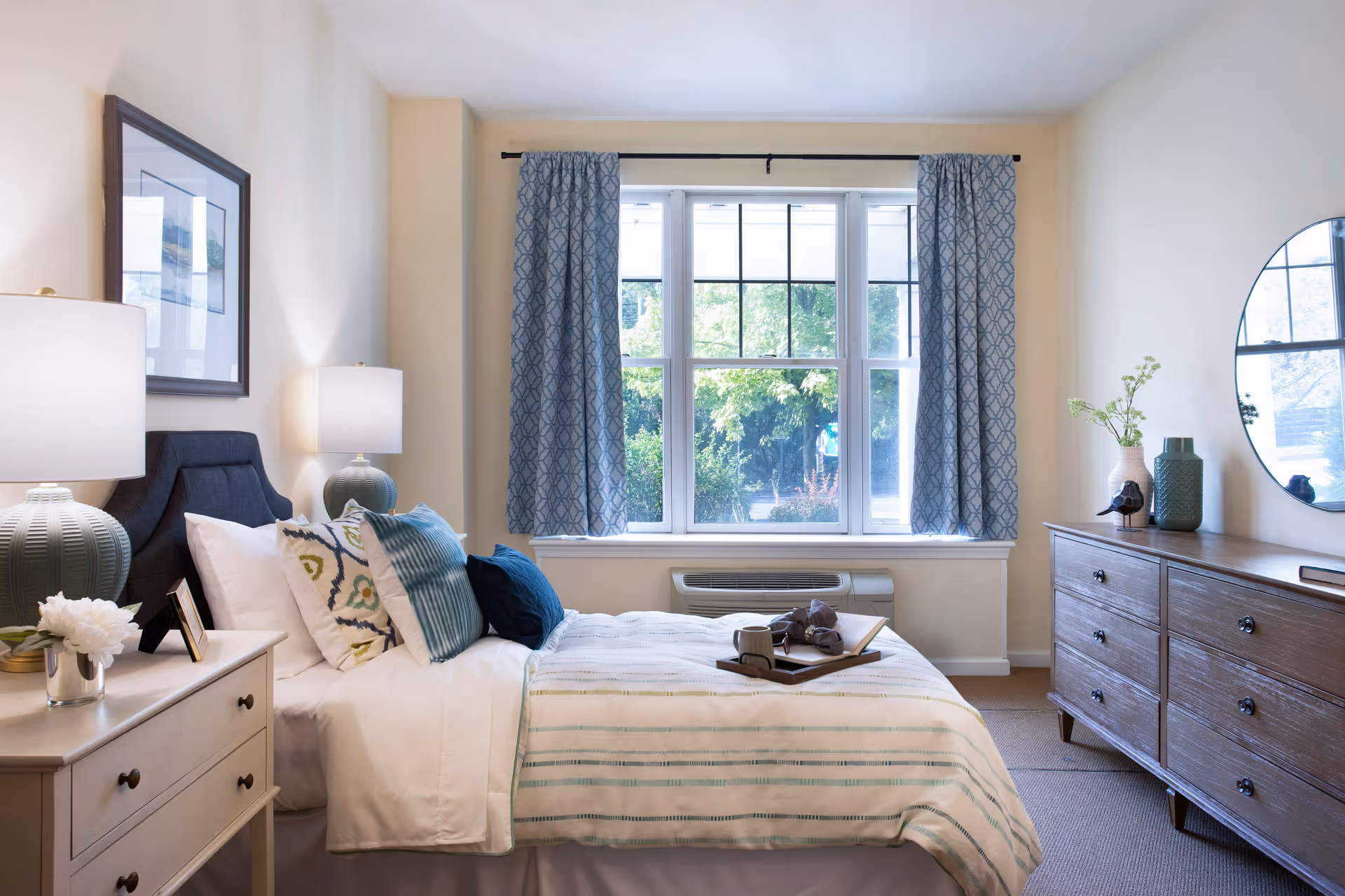 Sunlit furnished bedroom with a made bed, nightstand and lamp, dresser with mirror, and a large window framed by blue curtains.