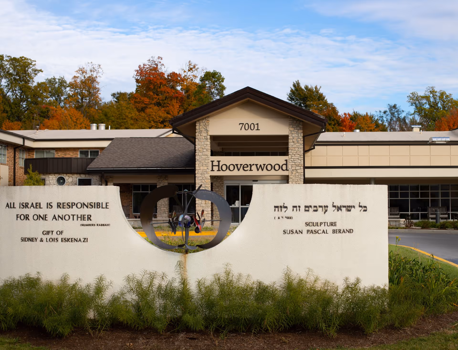 Entrance facade of the Hooverwood building (7001) with a front sculpture and a white inscription wall reading "All Israel is responsible for one another", set against autumn trees.