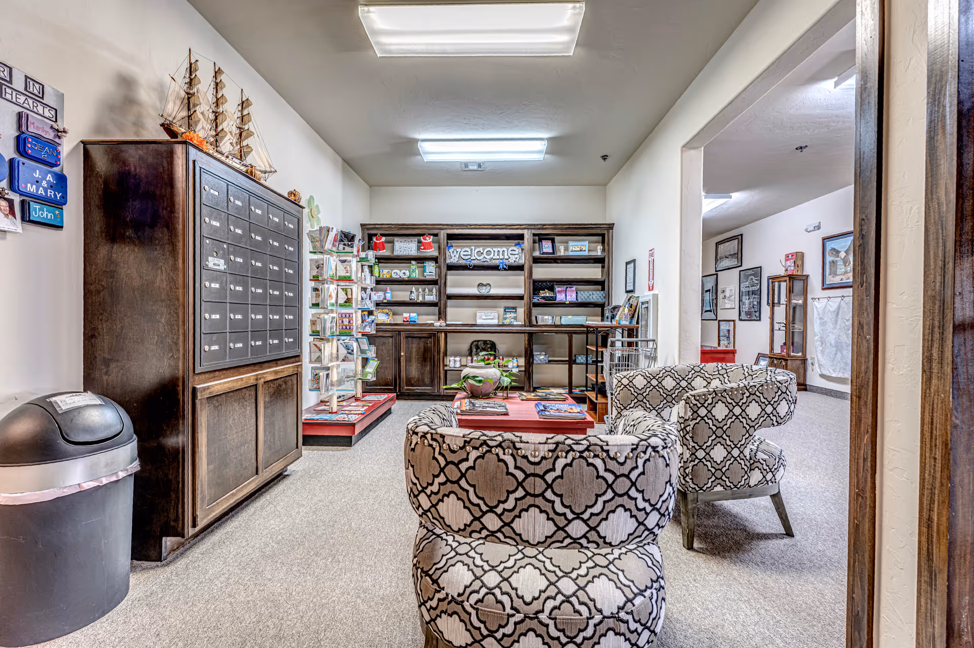Interior view of a senior living facility common area with two patterned armchairs facing a low table. To the left, there is a large wooden mailbox unit and a trash can. At the back, shelves hold various items including a welcome sign and greeting cards. The room has beige walls and carpeted floors with fluorescent ceiling lights.