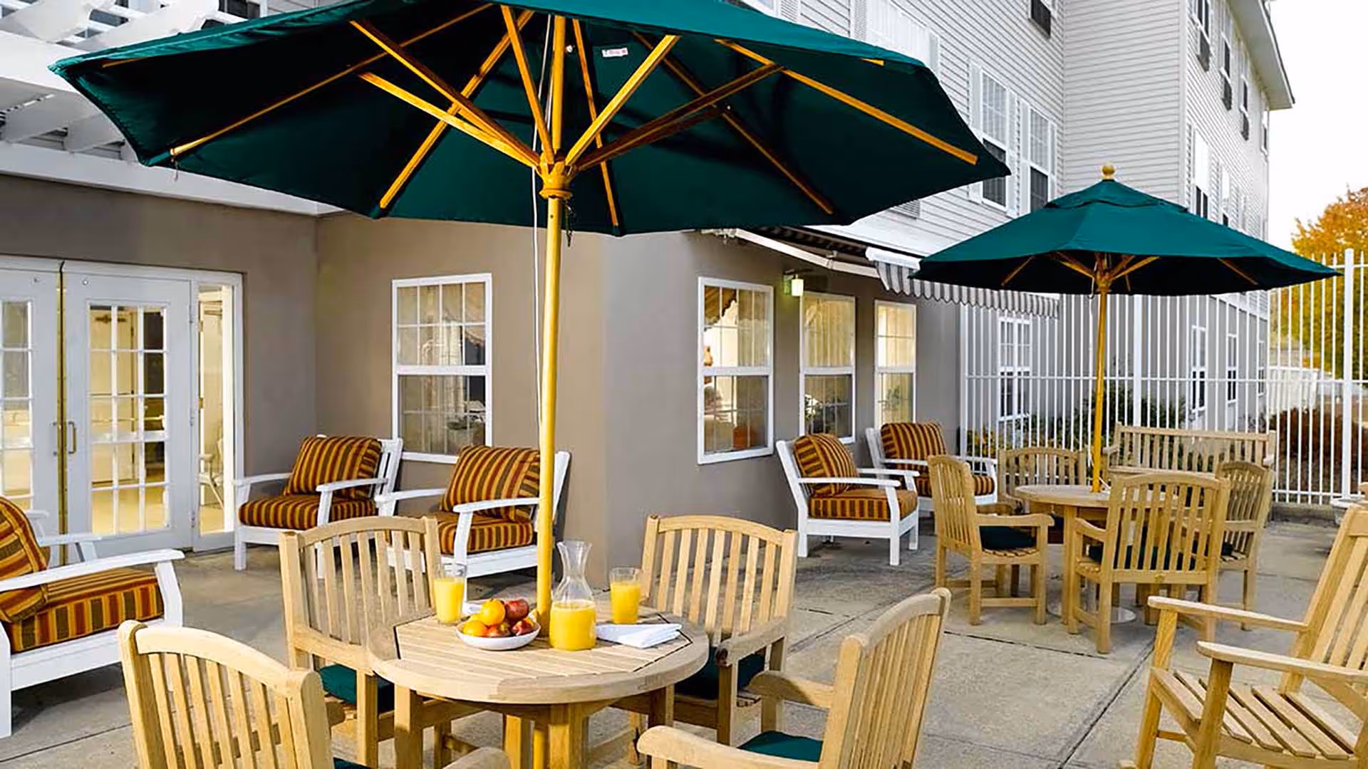 Outdoor patio area with wooden tables and chairs, each table shaded by a large green umbrella. Cushioned armchairs with striped upholstery are arranged along the building wall with windows and glass doors in the background. A pitcher of orange juice, glasses, and a bowl of fruit are on one of the tables.