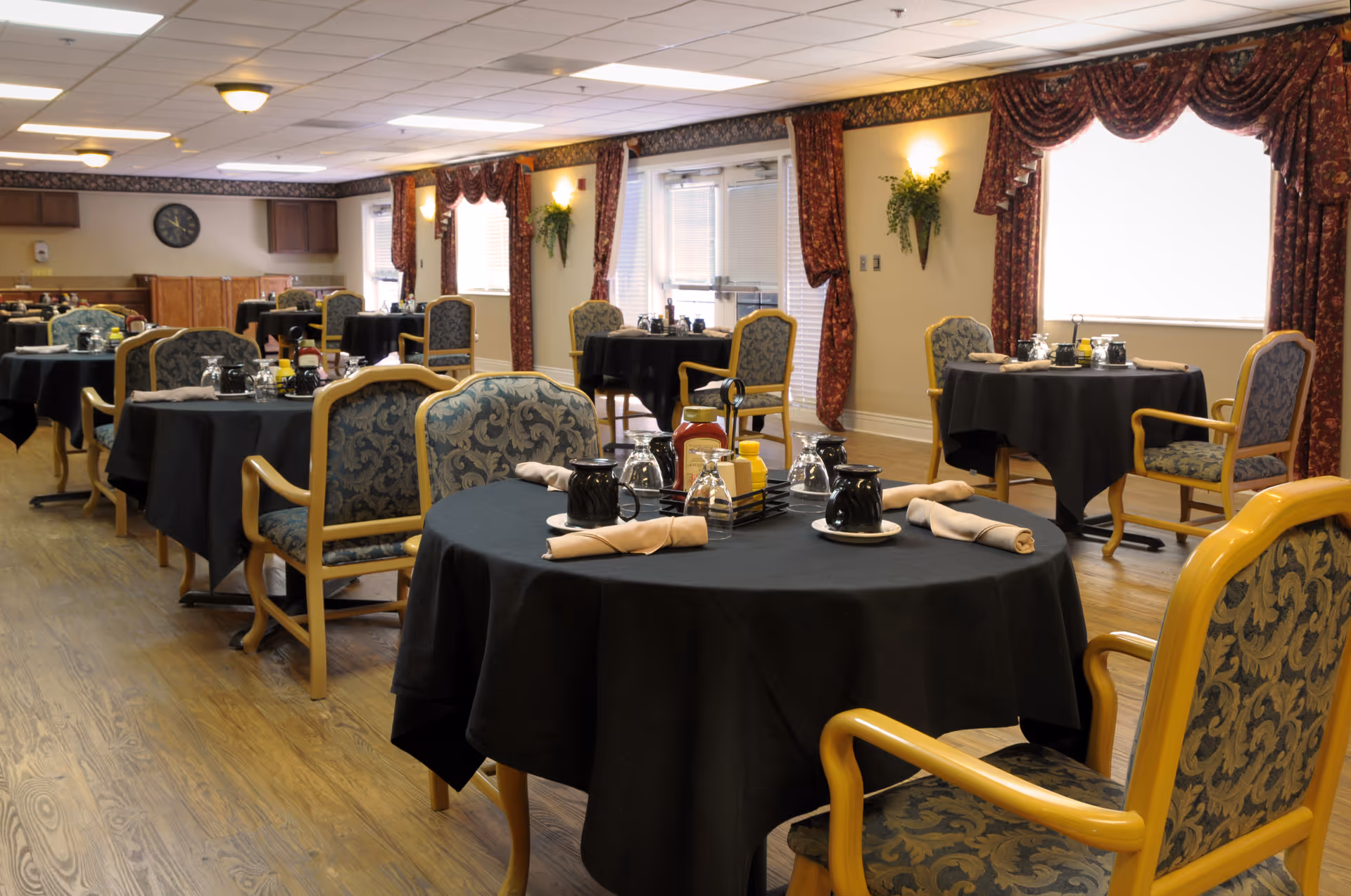 A dining room in a senior living facility with round tables covered in black tablecloths, each set with napkins, cups, and condiments. The room has wooden floors, patterned upholstered chairs, large windows with red curtains, and soft lighting from ceiling fixtures and wall sconces.