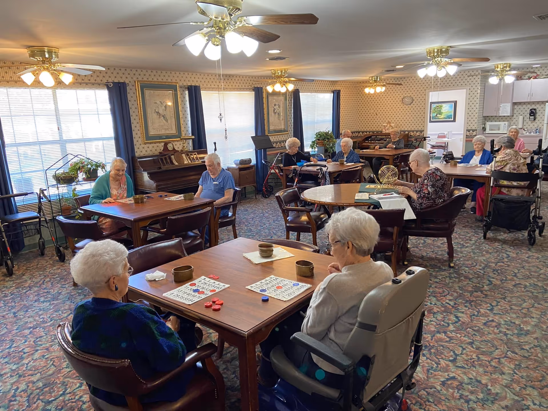 A group of elderly people sitting at tables in a well-lit room playing bingo. The room has patterned wallpaper, carpeted floor, ceiling fans with lights, and large windows with blue curtains. There is a piano against one wall and a kitchenette area in the background.
