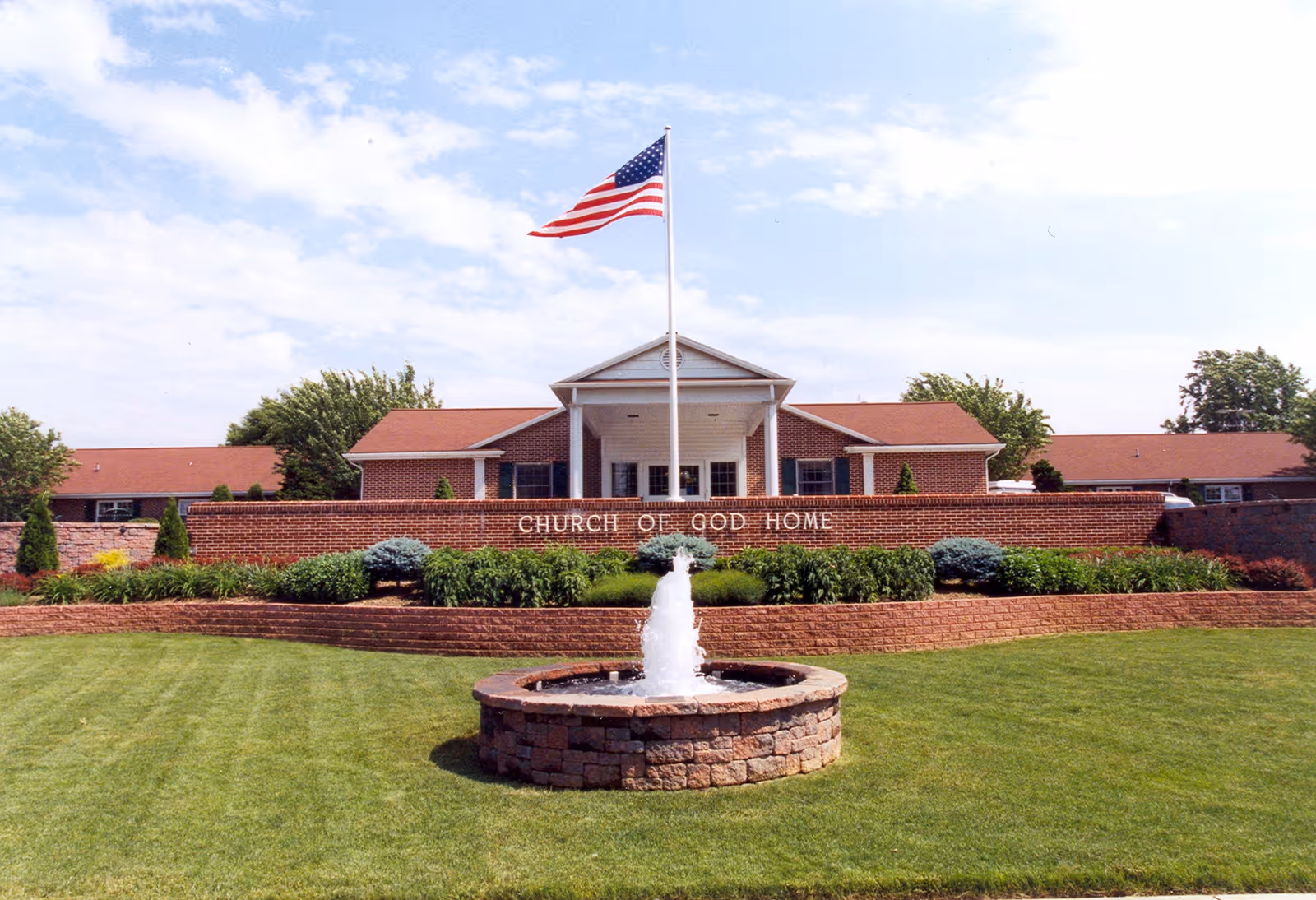 Front exterior view of the Church of God Home building with a brick facade, a central entrance with white columns, an American flag on a flagpole, a water fountain in the foreground, and well-maintained landscaping with green grass and shrubs.