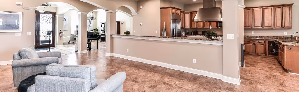 Spacious interior area with a seating space featuring two gray armchairs in the foreground, a kitchen with wooden cabinets and stainless steel appliances to the right, and an open entryway with arches leading to another room with a piano and chandelier in the background.