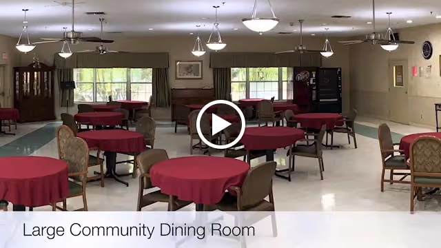 Large community dining room with multiple round tables covered with red tablecloths and surrounded by chairs. The room has large windows with green curtains, ceiling lights, and a vending machine in the back corner.