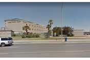 Exterior view of a large, beige multi-story building with a flat roof and minimal windows, situated behind a fenced area with some palm trees and a grassy lawn. A white SUV is parked on the street in front of the building, and a blue mailbox is visible on the sidewalk.