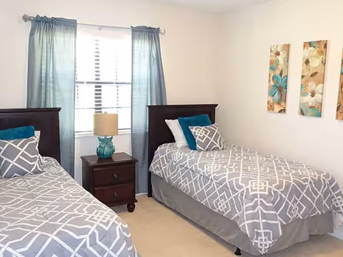 A bedroom with two twin beds featuring gray and white geometric patterned bedding, each with decorative pillows. Between the beds is a dark wooden nightstand with a turquoise lamp. A window with blue curtains is centered on the back wall, and three floral-themed paintings hang on the right wall.