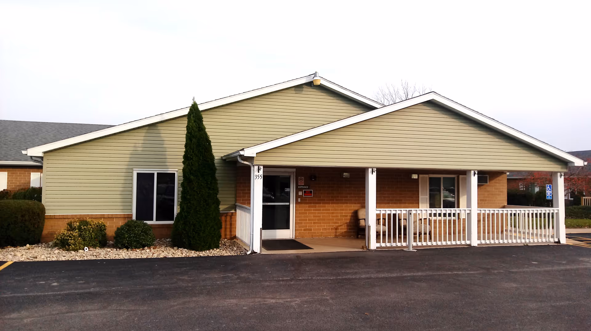 Exterior view of a single-story building with light green siding and brick lower walls. The building has a covered entrance with white pillars and railing, two chairs on the porch, and a small landscaped area with bushes and a tall evergreen tree. There is a parking lot in front with a visible handicap parking sign.