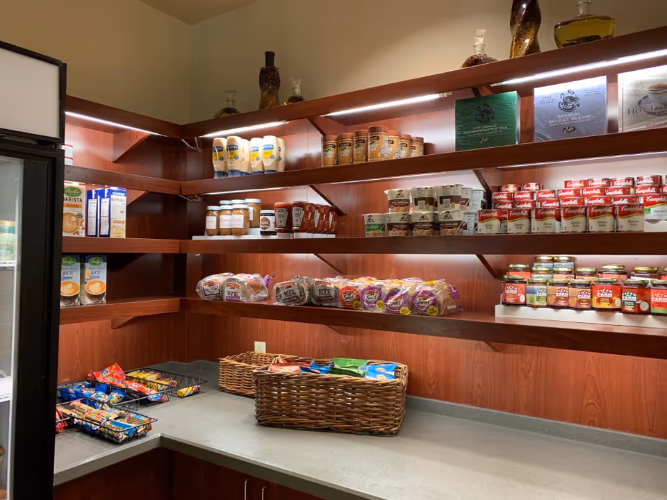 Shelves stocked with various food items including bread, canned soups, condiments, snacks, and tea in a pantry or small store area with wooden shelves and a countertop.