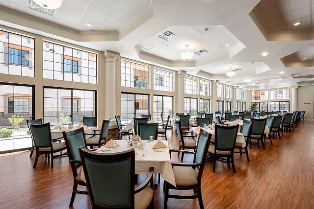 A spacious dining room with multiple tables covered in white tablecloths, each set with napkins, glasses, and silverware. The room features large windows allowing natural light to fill the space, wooden flooring, and green cushioned chairs arranged neatly around the tables.