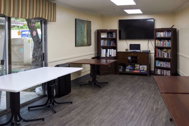 An interior common room with folding tables, bookshelves, a wall-mounted TV, and a sliding glass door to the outside.