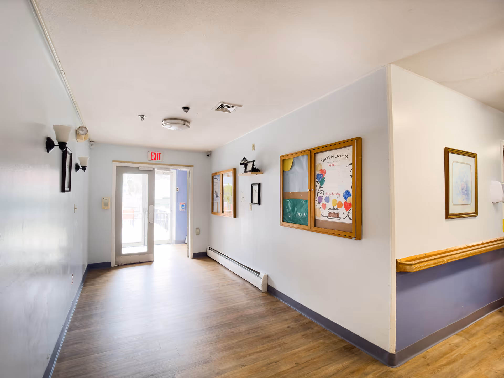 A well-lit hallway in Palmer Healthcare Center with light-colored walls and wooden flooring. On the right wall, there are bulletin boards displaying birthday announcements and other notices. The hallway leads to a glass door with an exit sign above it. Wall-mounted light fixtures and framed artwork are visible along the walls.